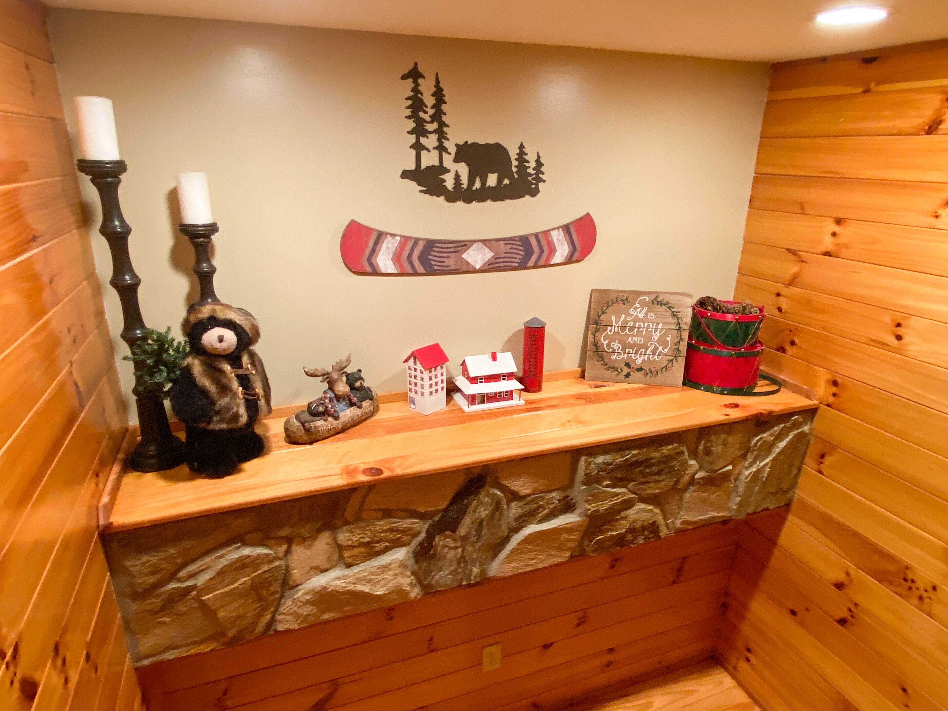 Interieur van een houten hutje met een stenen muur, een berenbeeldje, een elandbeeldje, een klein huismodel, een rode emmer en een bordje met de tekst 'It's Merry and Bright'.