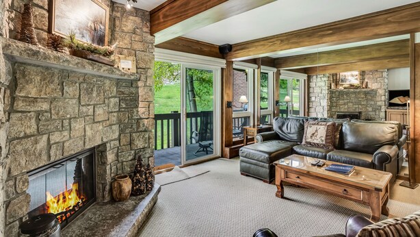 A cozy living room with a stone fireplace, leather sofa, and a large window overlooking a deck. - 🏔️Ski-In, Ski-Out Condo~Hot Tub~Interlude 205🏔️ (Snowmass Village)