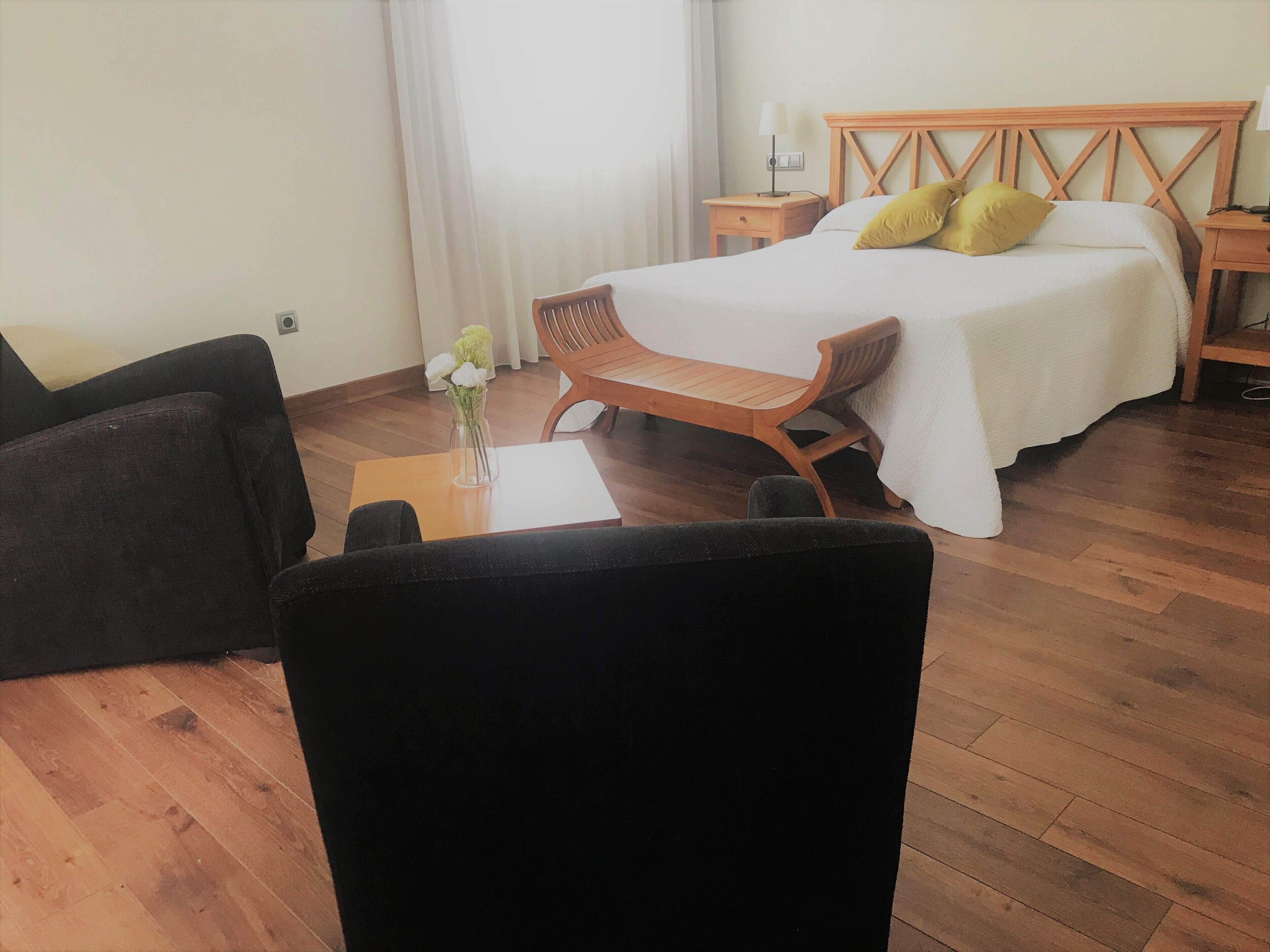 Habitación de hotel con una cama, una mesita de noche de madera, una silla, una pequeña mesa con un florero y una ventana con cortinas.