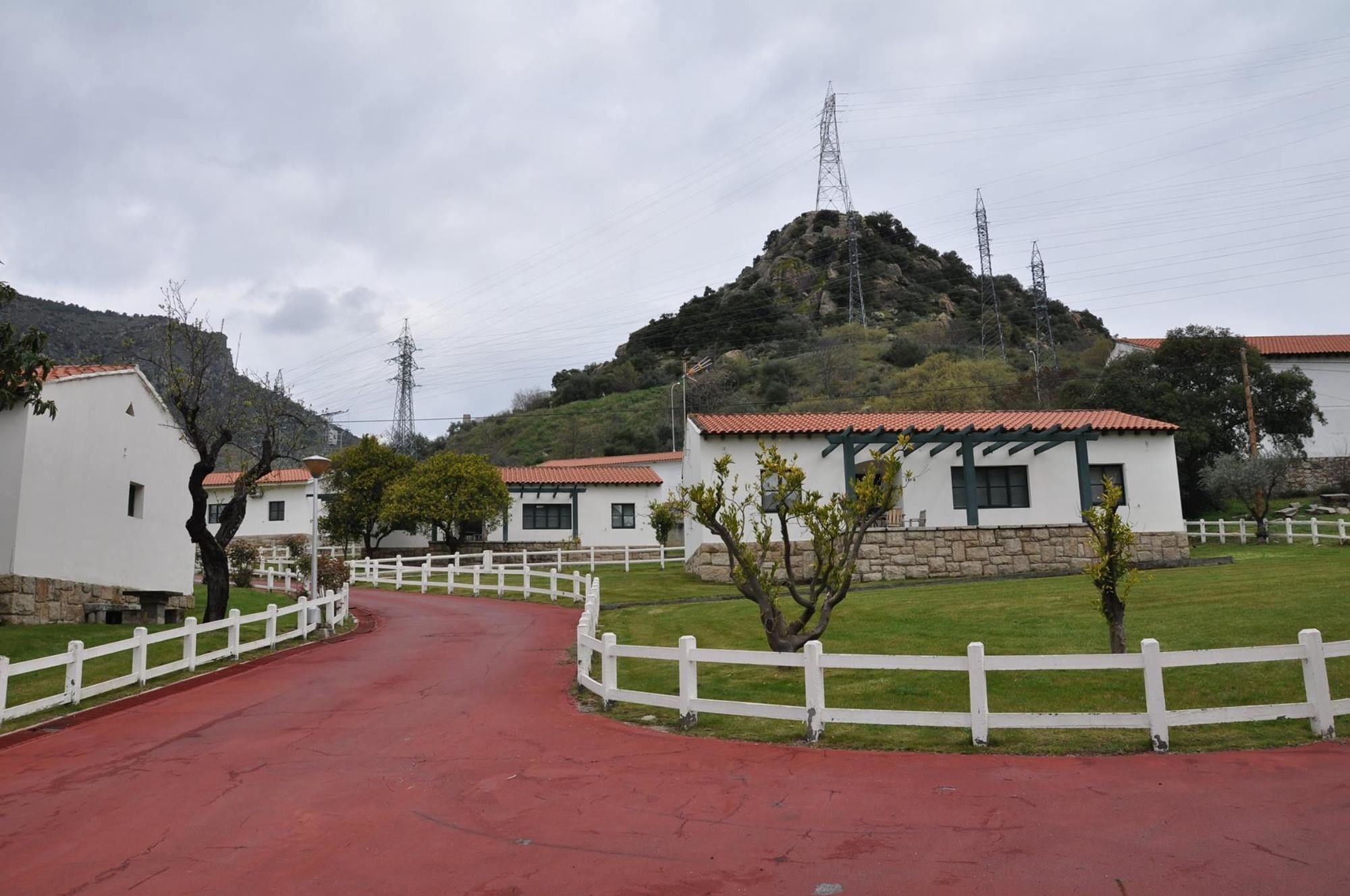 Un camino rural que conduce a edificios blancos con tejados rojos, rodeado de vegetación y una colina al fondo.