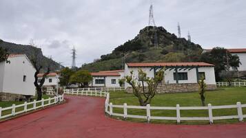 Un camino rural que conduce a edificios blancos con tejados rojos, rodeado de vegetación y una colina al fondo.