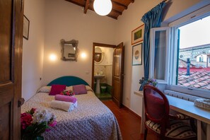 Un dormitorio con cama, una silla, escritorio, ventana y baño visible a través de una puerta abierta.