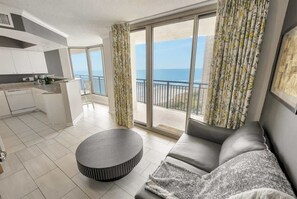 A modern living room with a grey sofa, a round coffee table, and a kitchen area with white cabinets. Large windows offer a view of the sea. - Beautiful Views, New 18th Floor Luxury, Brighton! (Myrtle Beach)