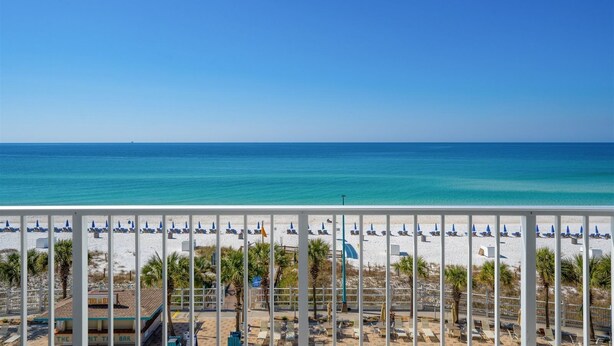Vista da praia com guarda-corpos brancos, guarda-sóis azuis e céu azul límpido.