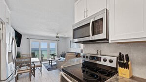 Uma cozinha moderna com micro-ondas, forno e fogão embutidos. Há uma área de jantar com mesa e cadeiras, e vista para o oceano através de grandes janelas.