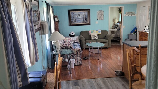 Sala de estar com sofá, uma mesinha e um espaço de jantar com cadeiras. Há uma janela com cortinas, um quadro na parede e uma porta que dá para outro cômodo.