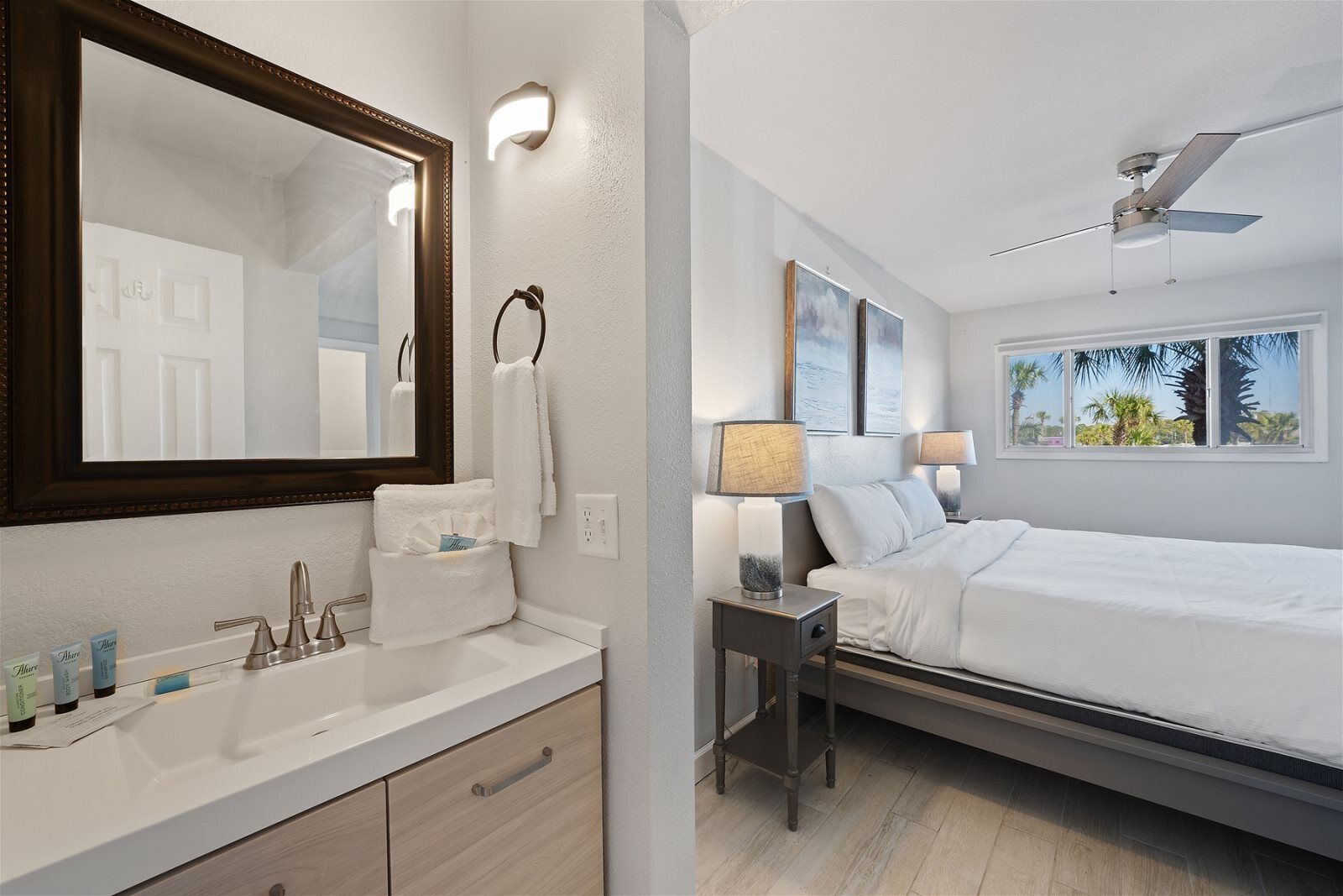 Une chambre d’hôtel avec un grand miroir, un lavabo blanc, une table de chevet avec une lampe, un lit et un ventilateur de plafond.