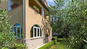 Un bâtiment à la façade beige, doté d’un socle en pierre et agrémenté d’un jardin bien entretenu, avec diverses plantes et une pelouse soigneusement taillée.