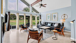 A spacious living room with a large window, a ceiling fan, a black leather sofa, wooden chairs, and a coffee table.