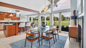 A modern kitchen with wooden cabinets, a glass dining table, and chairs. There is a large window with a view of a golf course.