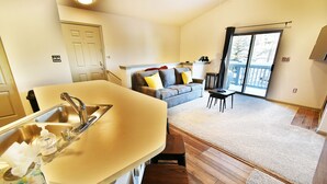 A compact kitchen with a sink and countertop, a dining area with a table and chairs, a sofa with cushions, and a sliding glass door leading to a balcony. - Walk to Lake Rec Path! Corner Unit Close to Everything! 2 Hot Tubs (Frisco)