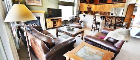 Sala de estar con sofás de cuero, una mesa de centro, televisor y una zona de cocina con alacenas de madera y refrigerador.