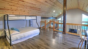Una cama litera con colchón y almohadas, suelo de madera, escalera con barandilla y vistas al exterior a través de amplios ventanales.