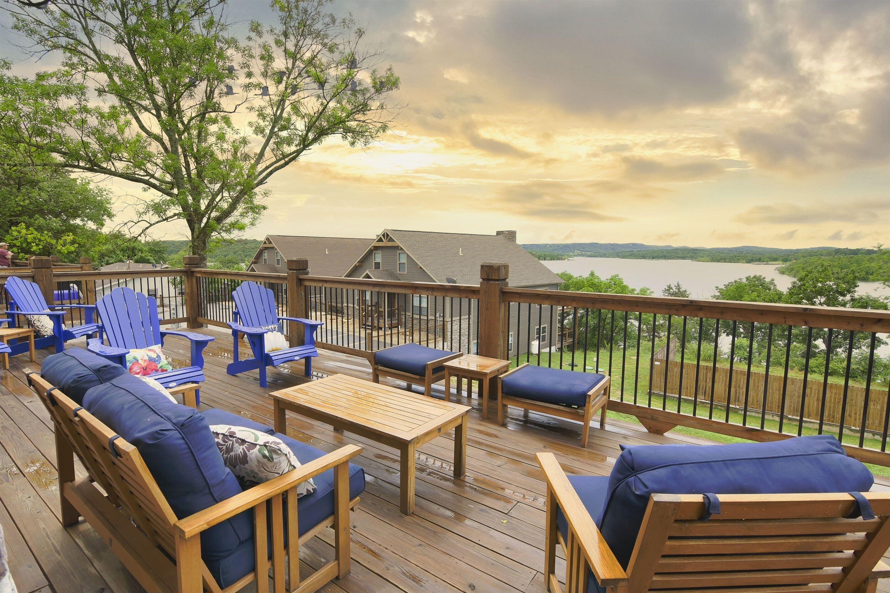 Eine Holzterrasse mit blauen Adirondack-Stühlen und einem Tisch, mit Blick auf einen See und Hügel.