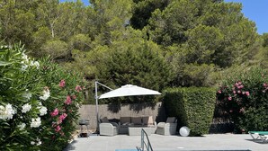 A swimming pool with a metal ladder, surrounded by a tiled patio. There are wicker lounge chairs and a white umbrella in the background.