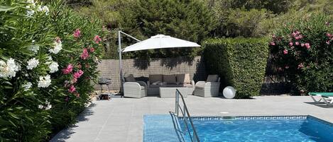 A swimming pool with a metal ladder, surrounded by a tiled patio. There are wicker lounge chairs and a white umbrella in the background.