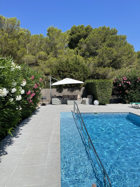 A swimming pool with a metal ladder, surrounded by a tiled patio. There are wicker lounge chairs and a white umbrella in the background. - Villa Honeysuckle (Son Parc)