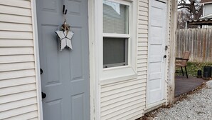 Una casa con puerta gris, ventana con persianas y una terraza de madera con grava.