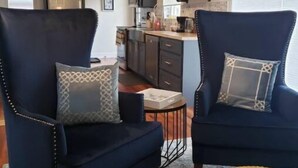 Una sala moderna con dos sillones azul oscuro, una mesita redonda y un taburete tejido. Se ve una cocina al fondo con armarios oscuros y una lámpara colgante.