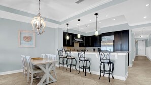 A modern kitchen with a central island, pendant lighting, and a dining area with a wooden table and chairs. - Escapes to the Shores 1702 by Liquid Life (Orange Beach)
