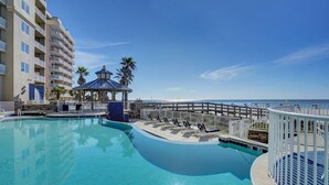 A swimming pool with lounge chairs and a gazebo, surrounded by a fence, with a beach and ocean view in the background. - Escapes to the Shores 1702 by Liquid Life (Orange Beach)