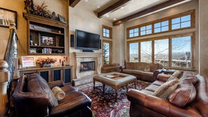 A spacious living room with leather sofas, a fireplace, a television, and large windows offering a view of the outdoors.