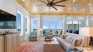 A spacious living room with a wooden ceiling, large windows, a flat-screen TV, a sofa, a coffee table, and a view of the ocean. - Seaside East and West ( 2 Properties - 24 Bedrooms) (Gulf Shores)