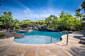Una piscina con cubierta de piedra, rodeada de vegetación tropical y un cielo azul despejado.