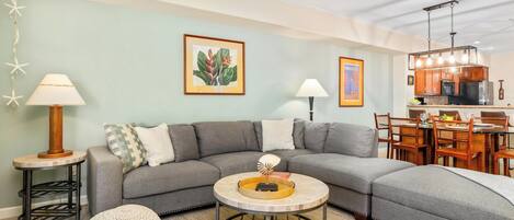A modern living room with a gray sectional sofa, a round coffee table, a small side table, and a dining area with wooden chairs and a table.