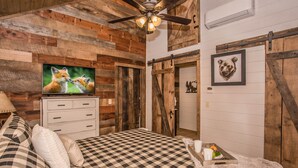Une chambre rustique avec une couvre-lit à carreaux, des murs en bois, un ventilateur de plafond, une télévision et une tête d’animal encadrée accrochée au mur.