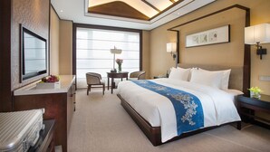 A hotel room with a large bed, a desk with a flat-screen TV, a chair, a small table with a lamp, and a window with blinds. - Kingdom Narada Grand Hotel Yiwu (Jinhua)