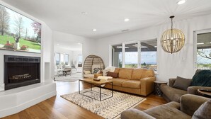 A spacious living room with a fireplace, a large window offering a view of the outdoors, and a modern chandelier. - WOW! Luxury Wine Country Estate - 6 Bedrooms • Hot Tub • Game Room (Temecula)