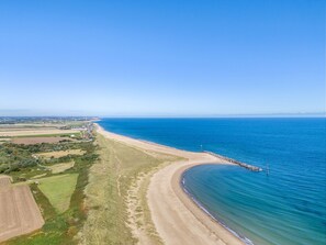A coastal landscape with a sandy beach, a clear blue sea, and a distant shoreline with buildings and fields.