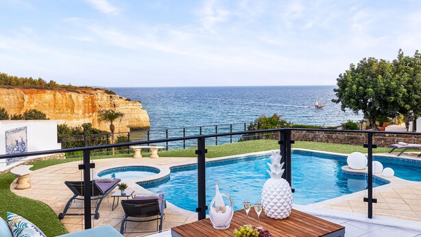A poolside area with a view of the sea, a wooden table with a pineapple, and blue cushioned seating. - Seafront View next to the famous “Benagil Caves” (Lagoa)