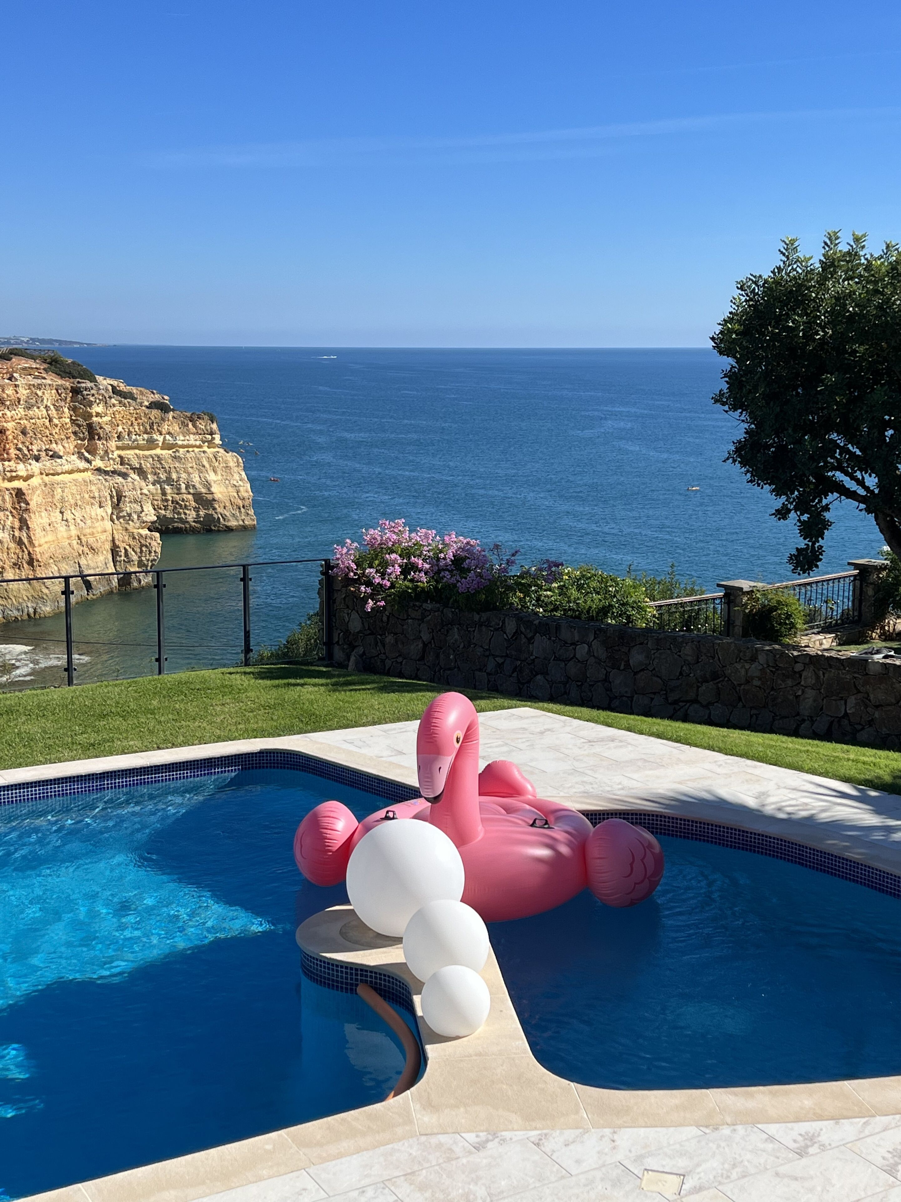 A swimming pool with a pink flamingo float, surrounded by a stone wall and a view of the ocean.