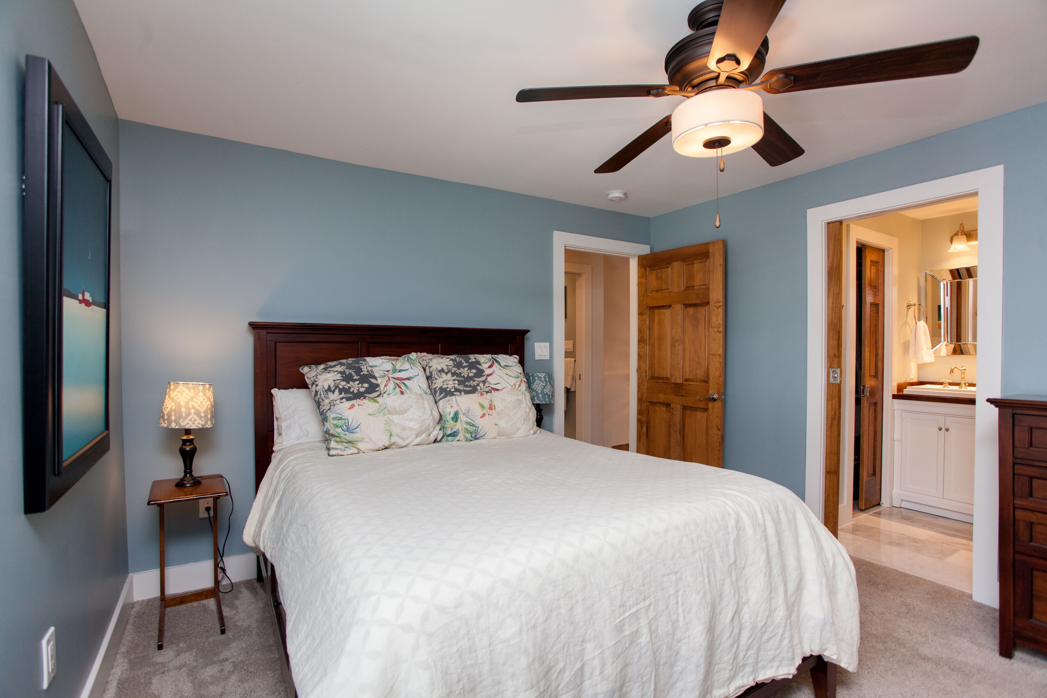 A bedroom with a bed, a ceiling fan, a nightstand with a lamp, and a door leading to a bathroom.