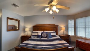 Une chambre à coucher avec un grand lit, deux tables de chevet avec des lampes, un ventilateur de plafond, une fenêtre avec des stores et une commode en bois.