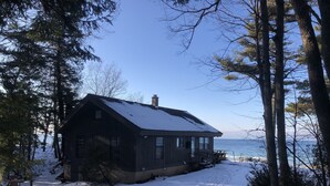 Eine Holzhütte mit schneebedecktem Dach, am Waldrand gelegen, mit Blick auf das Wasser.