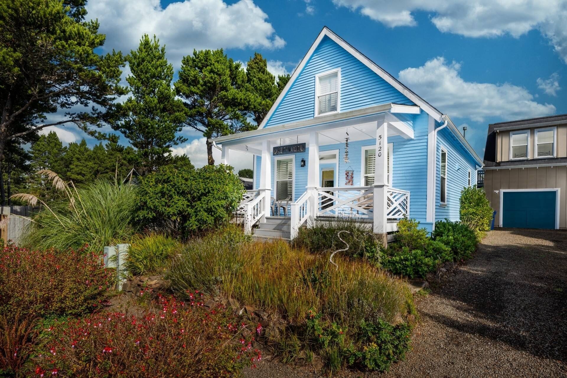 Uma casa de dois andares com fachada azul e detalhes brancos, cercada por vegetação exuberante e um caminho de acesso de brita.