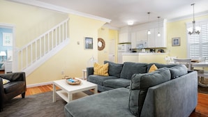 A living room with a gray sectional sofa, a white coffee table, and a gray ottoman. There is a staircase with white railings leading to an upper level. The kitchen is visible in the background with white cabinets and a white countertop. - Cozy Coastal Getaway, Open Floor Plan, Hot Tub, BBQ, Fenced Yard, Pet Friendly, Short Walk To Beach (Depoe Bay)