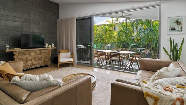 A modern living room with a TV, sofa, and dining area with a view of greenery. - Marie and Fred's Byron Villa (Byron Bay)