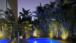 A well-lit outdoor pool area with a stone pathway leading to the pool, surrounded by tropical plants and a stone building. - Marie and Fred's Byron Villa (Byron Bay)