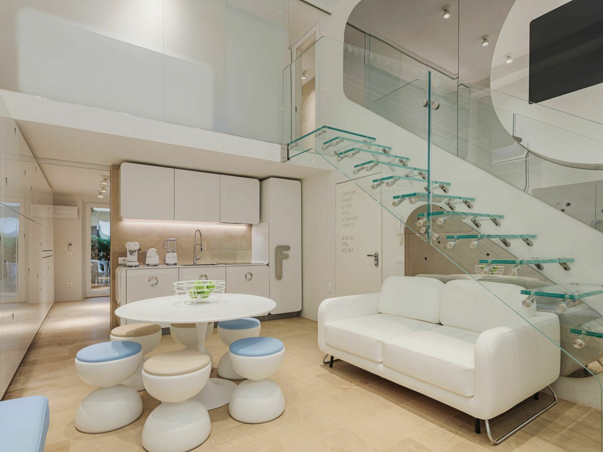 A modern interior with a white sofa, round table, and stools, a kitchen area, and a glass staircase.