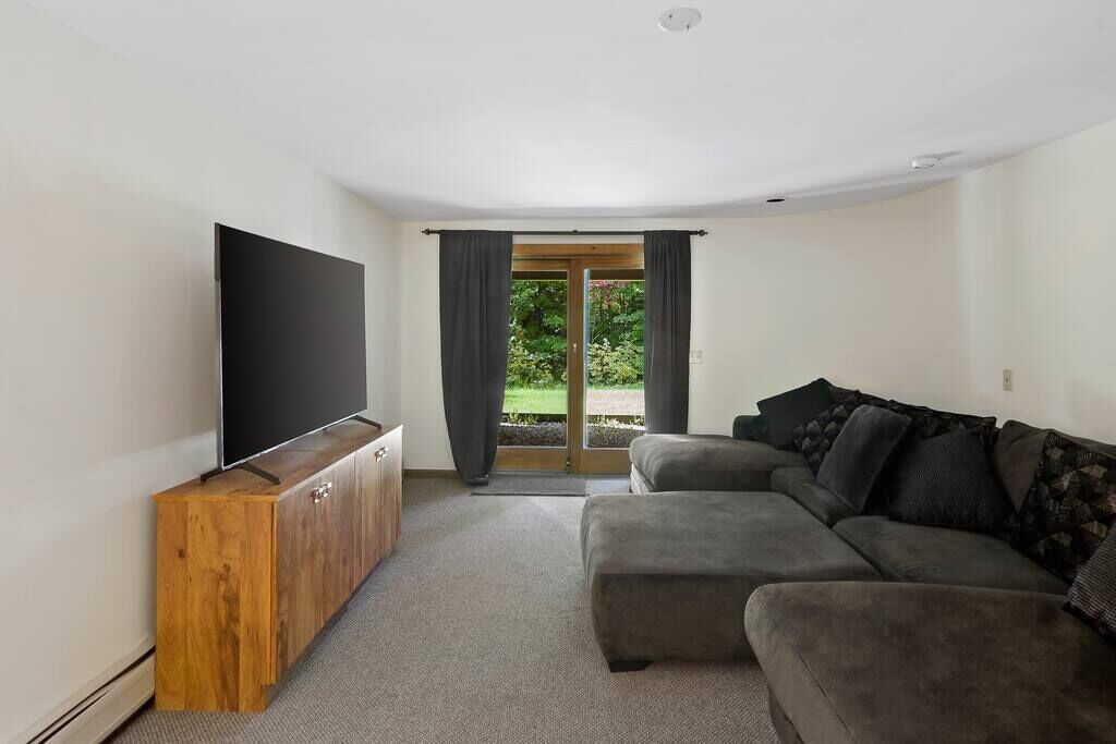 Una sala de estar con una televisión de pantalla plana sobre un mueble de madera, un sofá gris y un ventanal corredizo que da a un jardín.