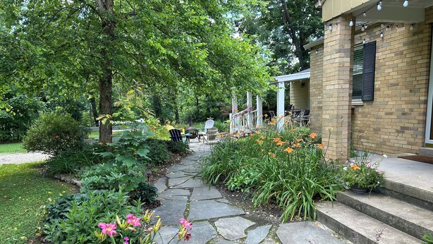 A stone pathway leading to a building with a brick facade, surrounded by lush greenery and flowering plants.