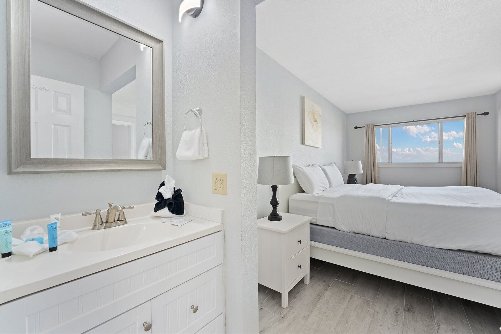 A hotel room with a large mirror, a white vanity with a sink, a bed with white bedding, and a window with a view of the sky.