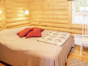 A log cabin bedroom with a bed, pillows, a patterned throw, a white chair, and a window.