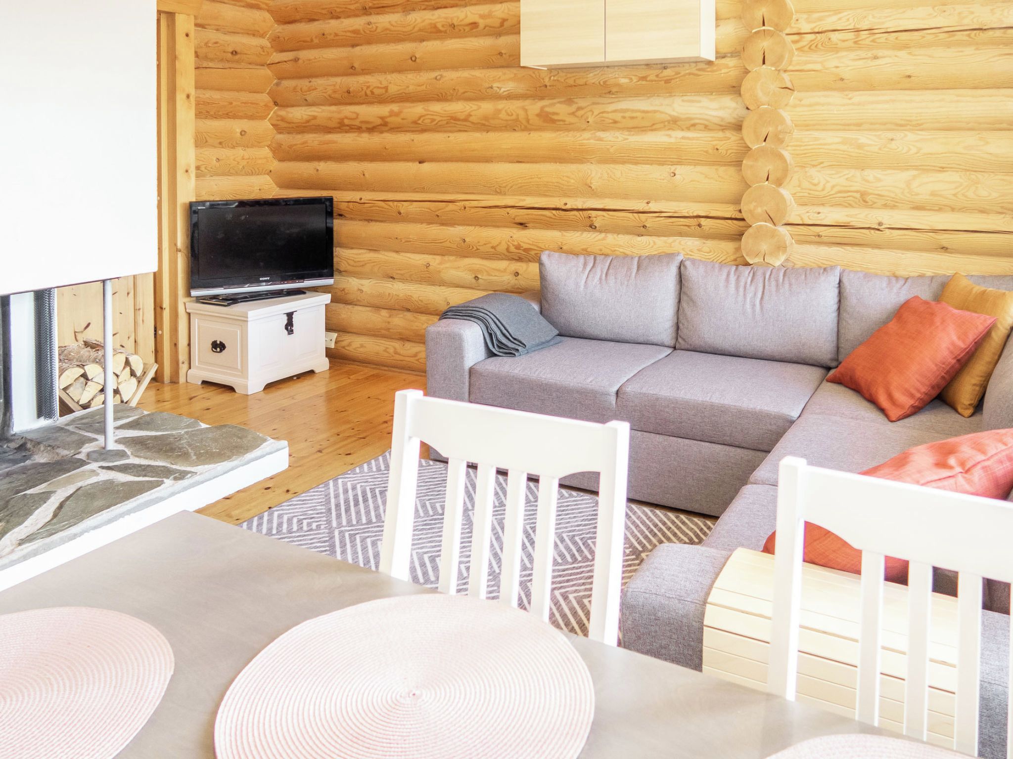 A cozy log cabin interior with a grey sofa, white chairs, a television, and a stone fireplace.