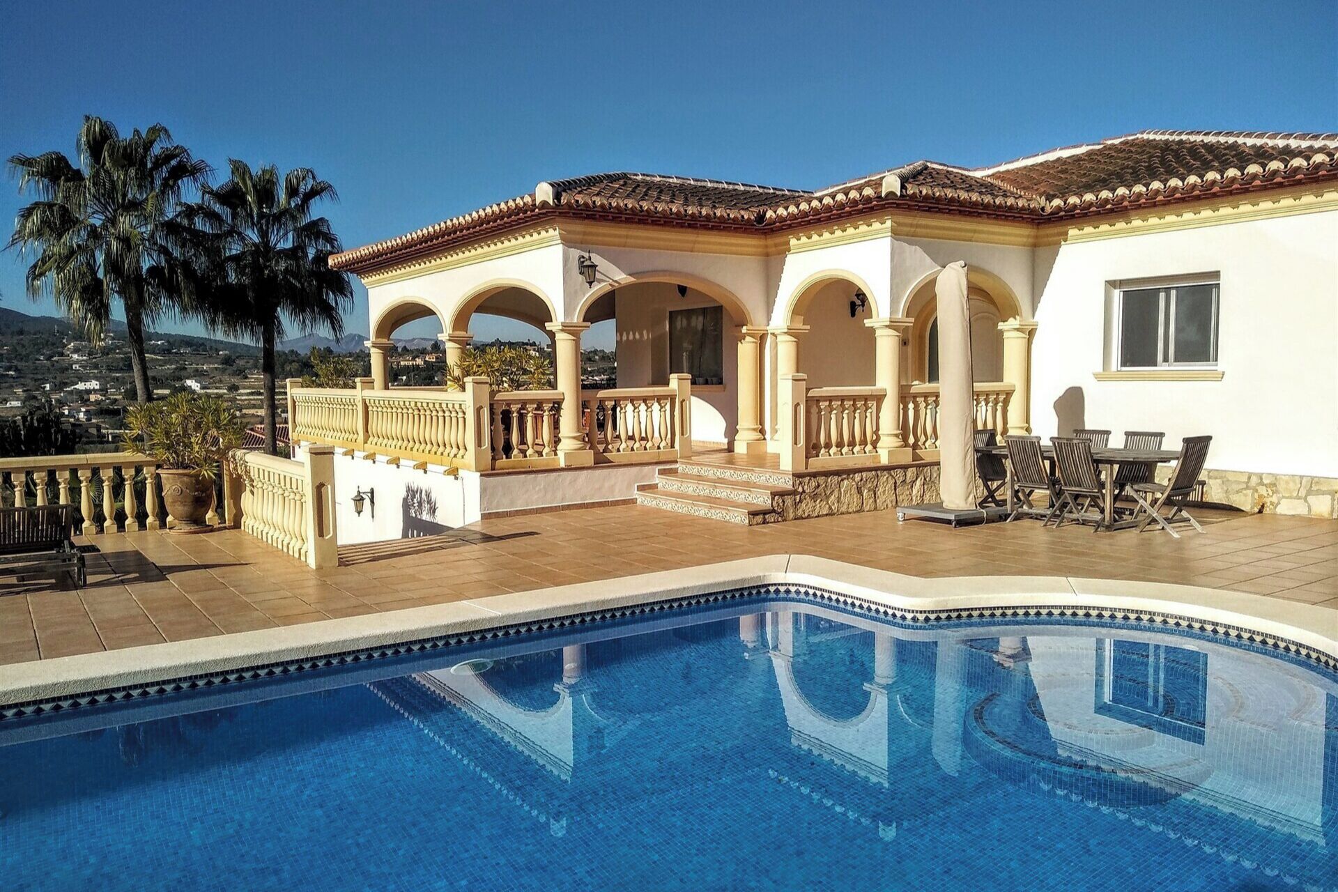 A poolside area with a clear view of a pool, a patio with outdoor furniture, and a building with arches and a tiled roof.