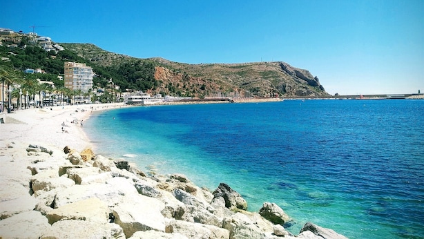 A coastal beach with clear blue waters, rocky shoreline, and buildings on a hillside. - Villa - Sleeps 7 - Pool - Parking (Javea)
